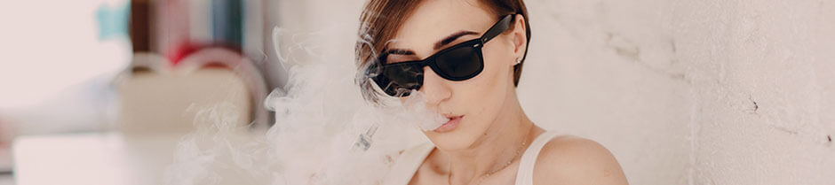 A pretty lady with dark glasses is exhaling vapor while holding a marijuana vape pen